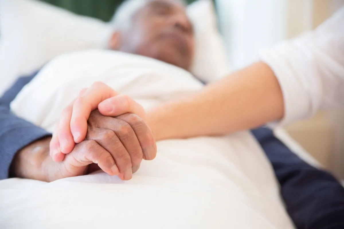 A companion gently holding the hand of an older man resting in bed