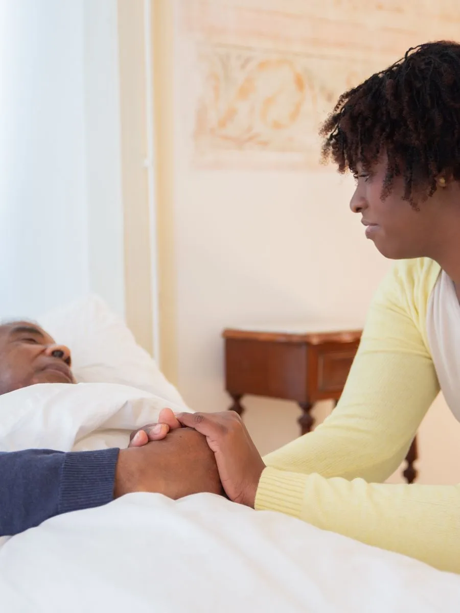 A young companion holding the hand of an older man resting in bed, a quiet moment of care at home