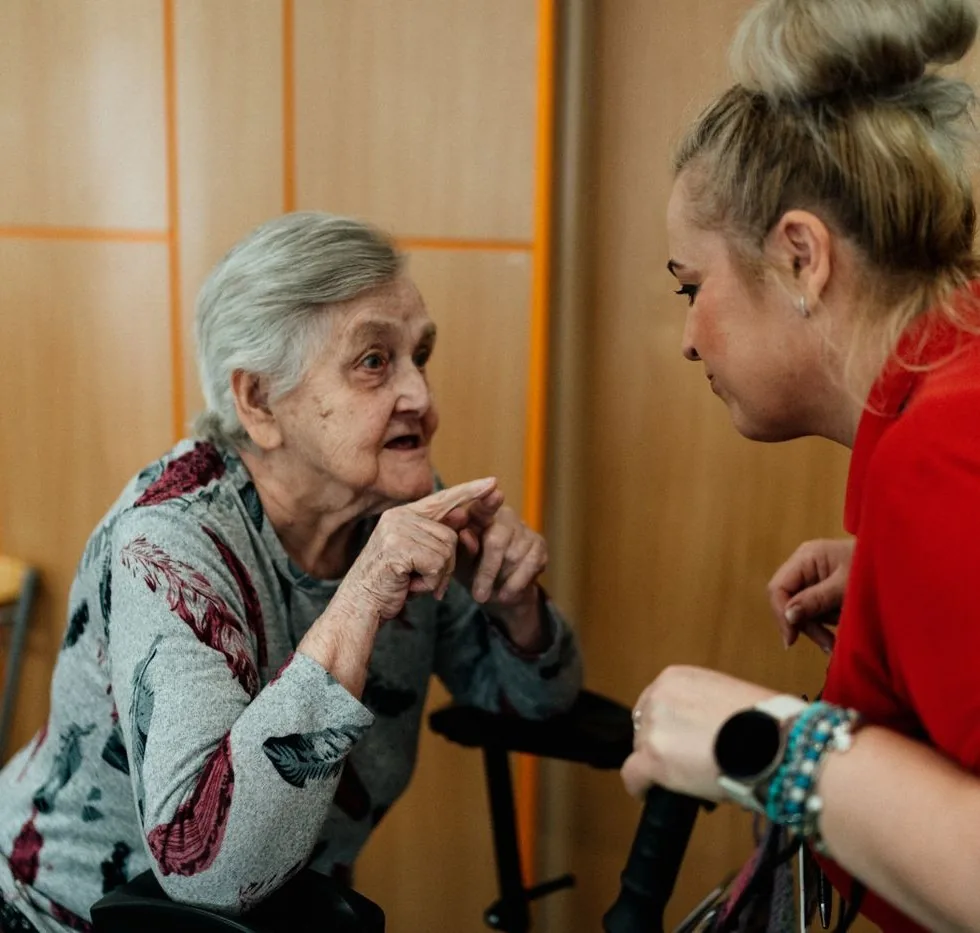 A caregiver leaning in at eye level, listening patiently as an older woman speaks and gestures in a bright room